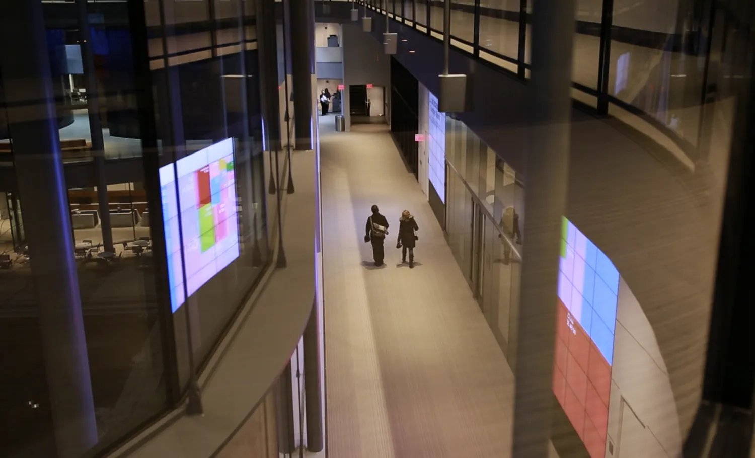People walking through the hallway at night-time beside large digital wall displays.