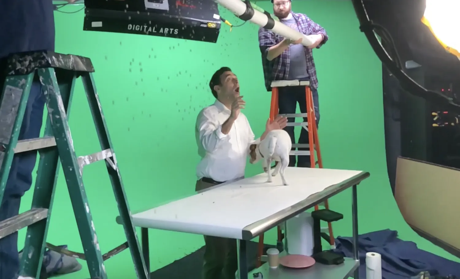 Video production crew filming a man and a dog on a vet's examination table in a green screen film set.