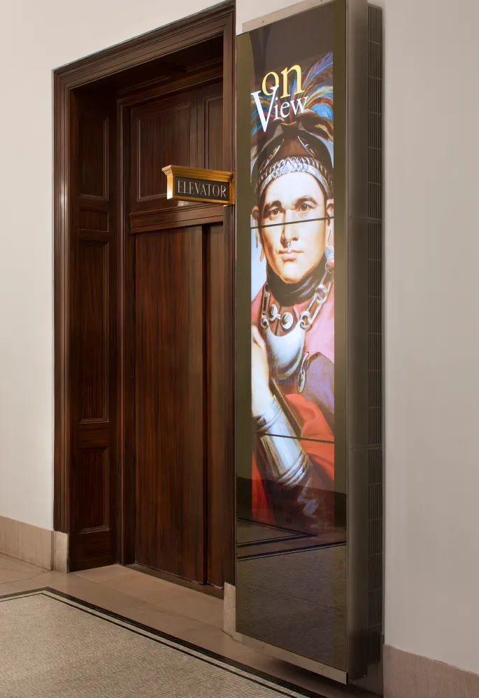 A vertical media display previewing ongoing exhibits, positioned in front of the museum's elevator.