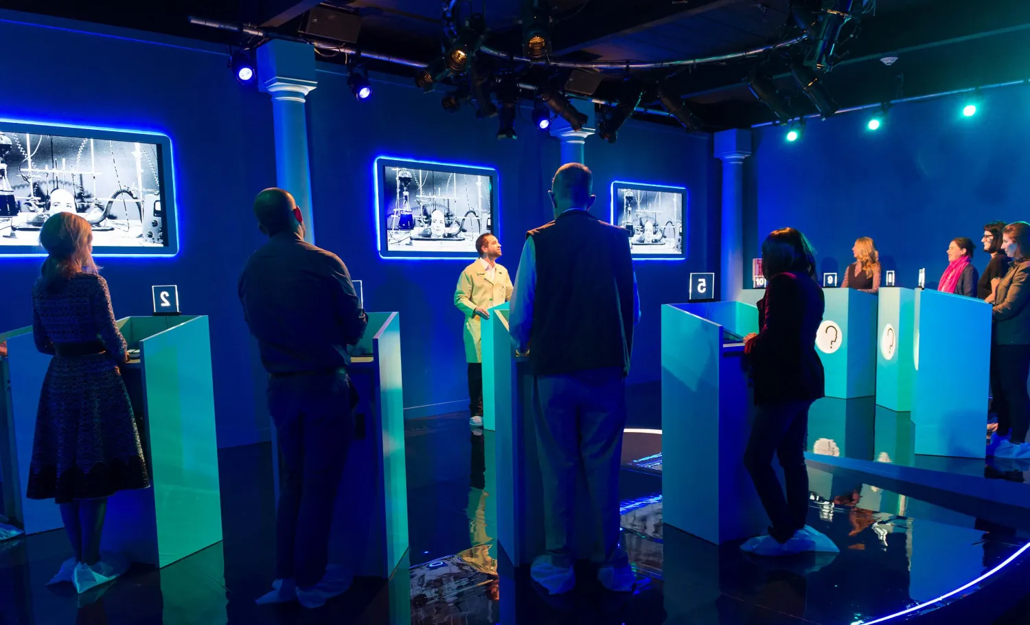 Group of people standing behind podiums listening while a host speaker stands in front of them talking.