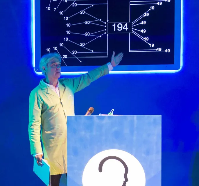 Host David Byrne talking behind a podium while presenting media on a digital blackboard screen.