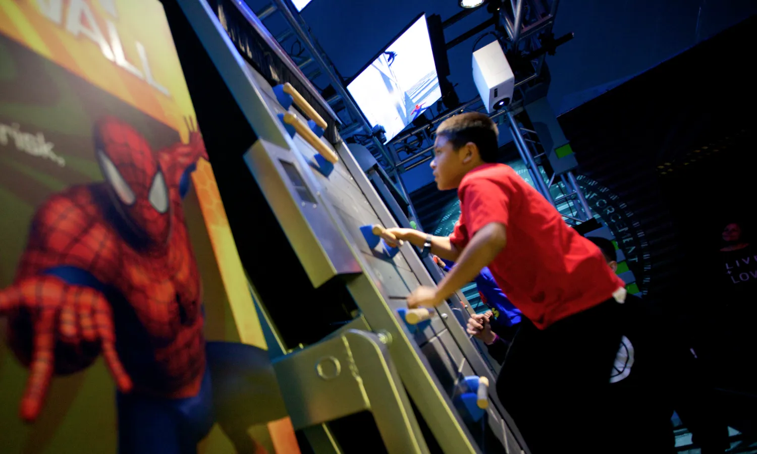 A child climbs a vertical wall while looking upwards towards a digital screen above beside a printed panel of Spider-Man.