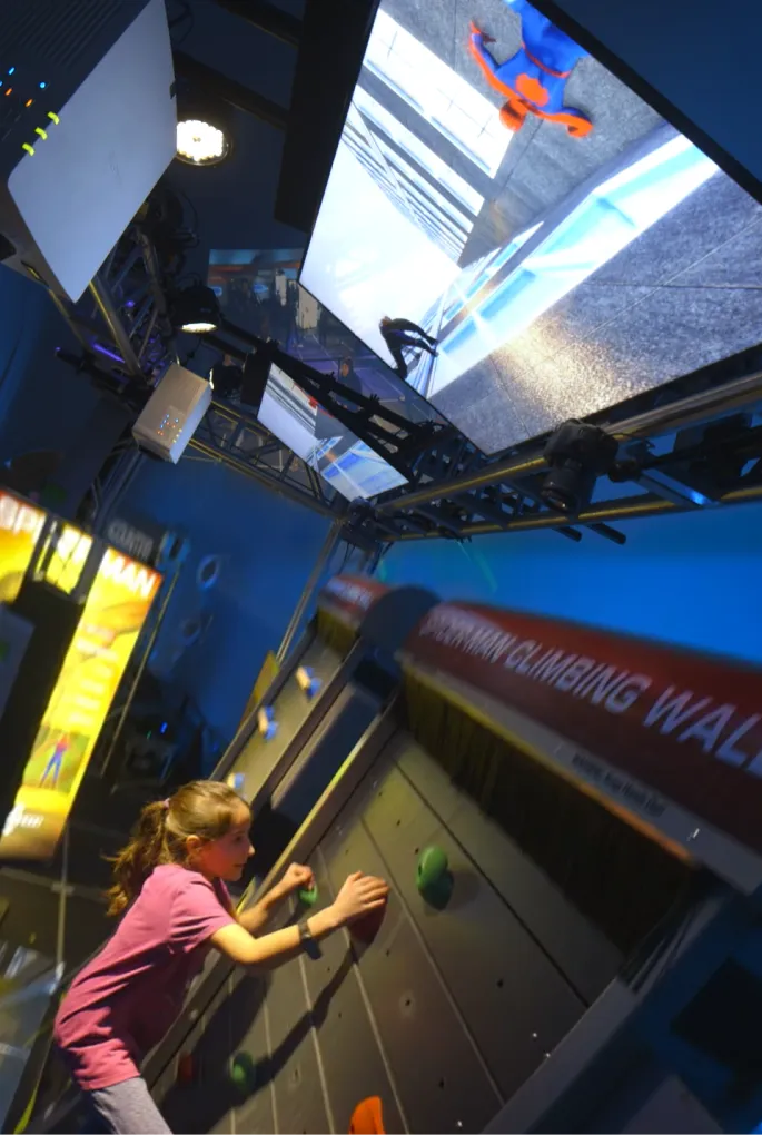 A girl climbs a rotating vertical wall while looking at an animated Spider-Man on the screen above.