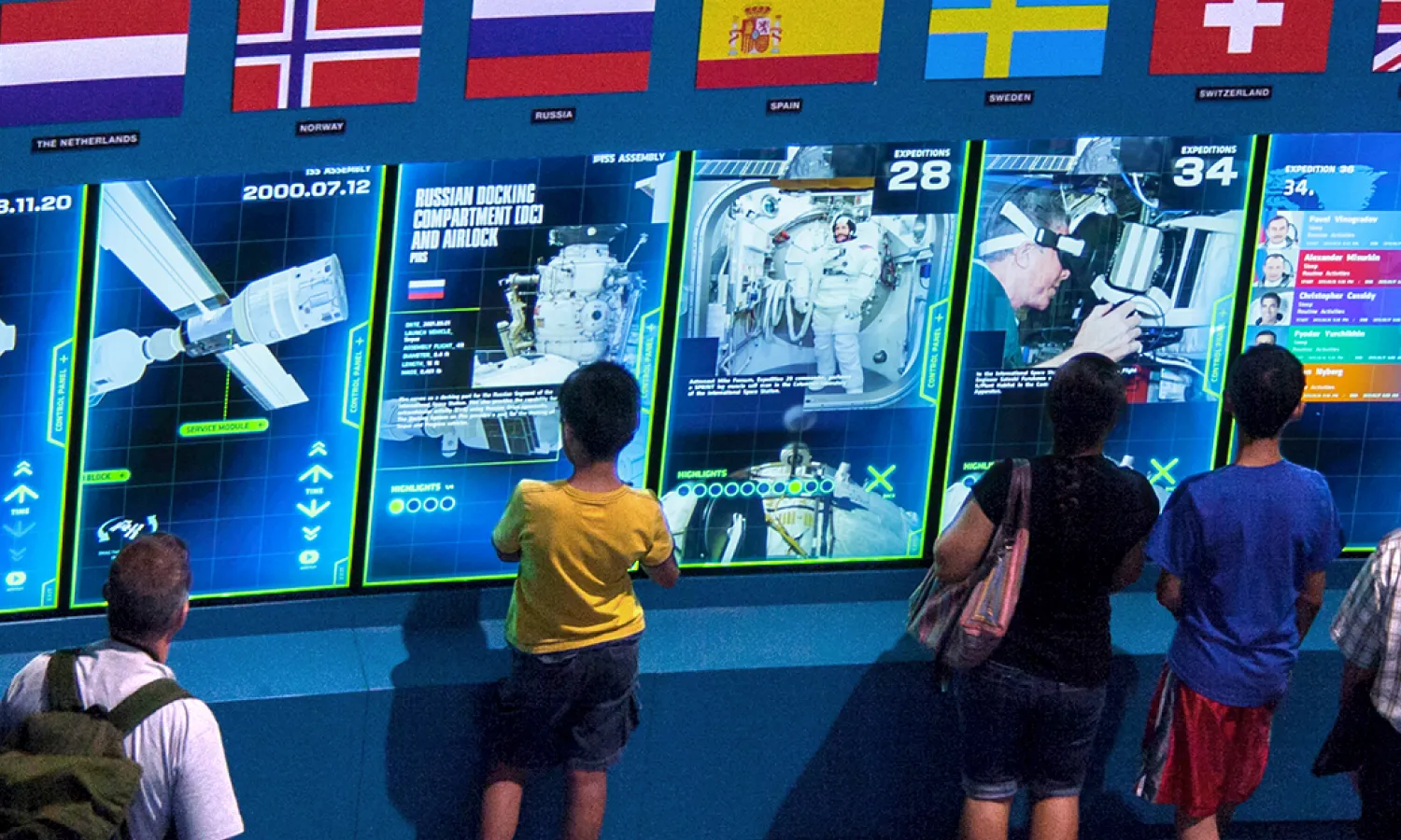 Multiple museum visitors standing in front of digital screens reading the exhibition text.