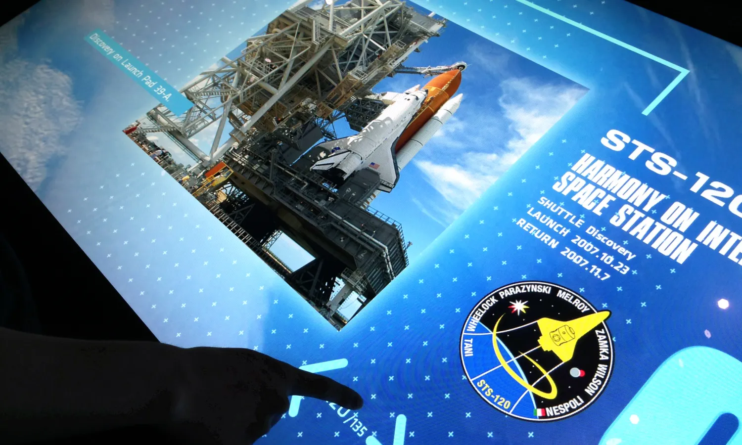 Visitor's hand interacting with a digital touchscreen viewing content on space shuttle.